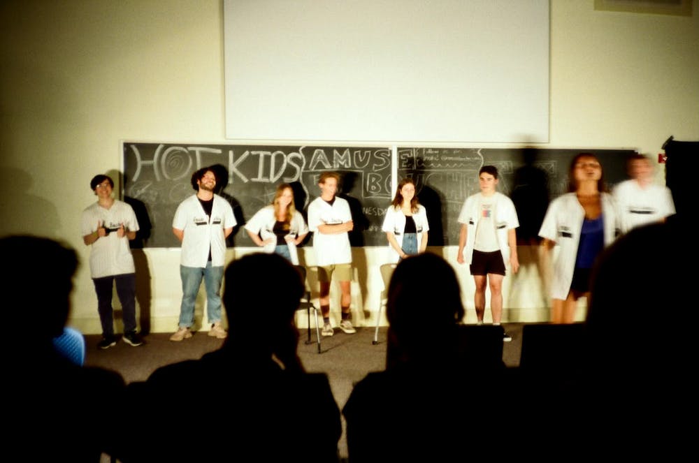 The members of Amuse Bouche wear baseball jerseys and stand in front of a chalk board, facing an audience.
