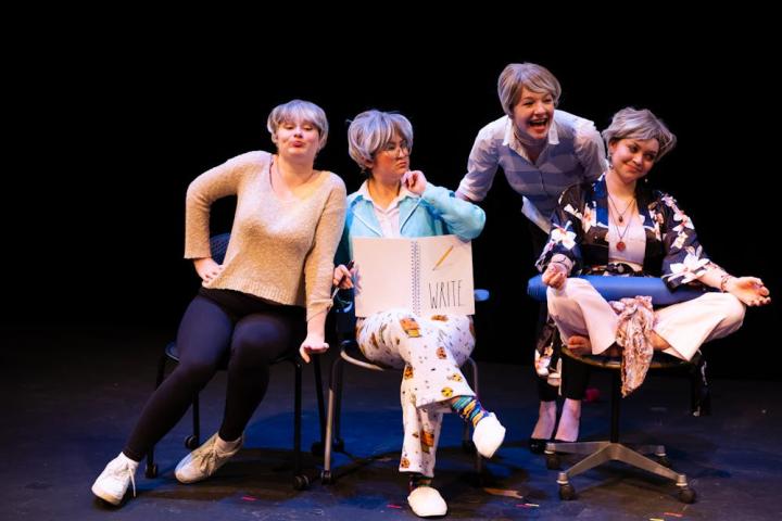 Four students sit in rolling chairs onstage, each wearing a short gray wig. 