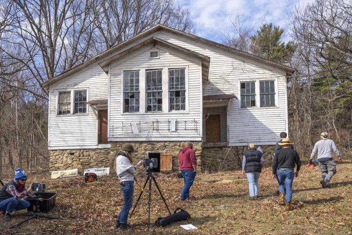 Seven people stand in the woods in front of a white building in disrepair. 