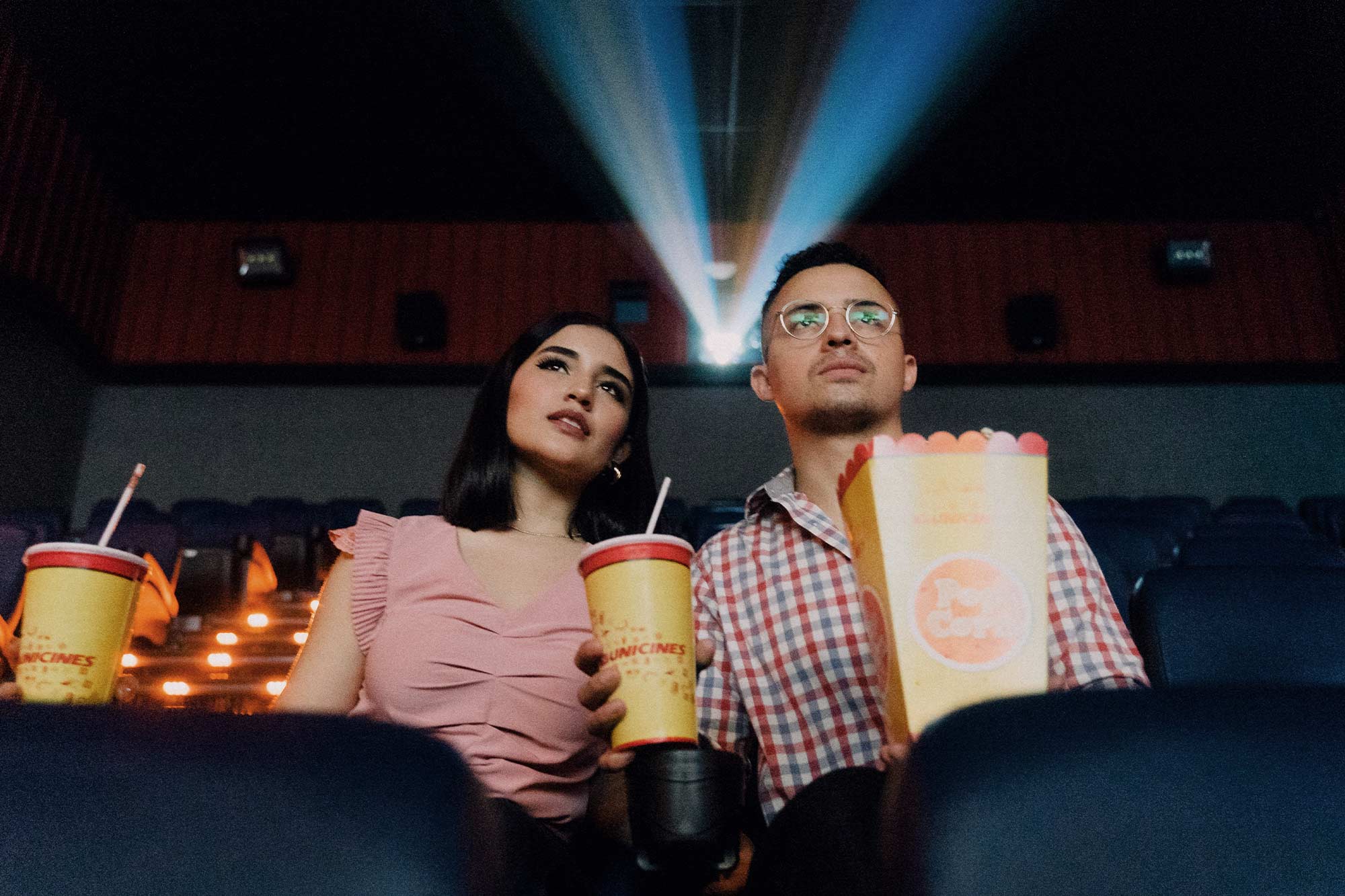 A man and woman sit side-by-side in a movie theatre, with popcorn and drinks