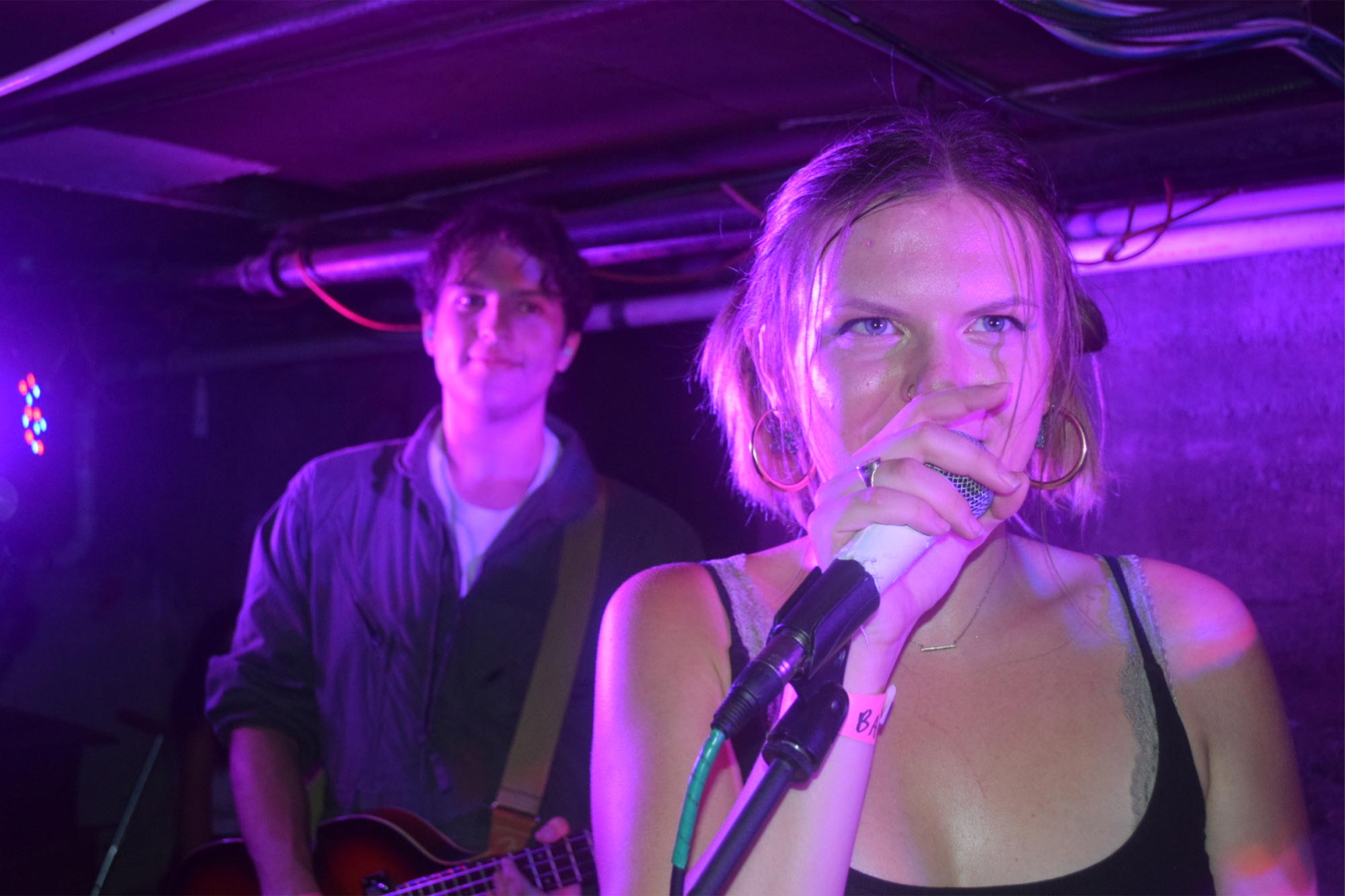 A photo of a person sining in the foreground while someone plays guitar in the back. 