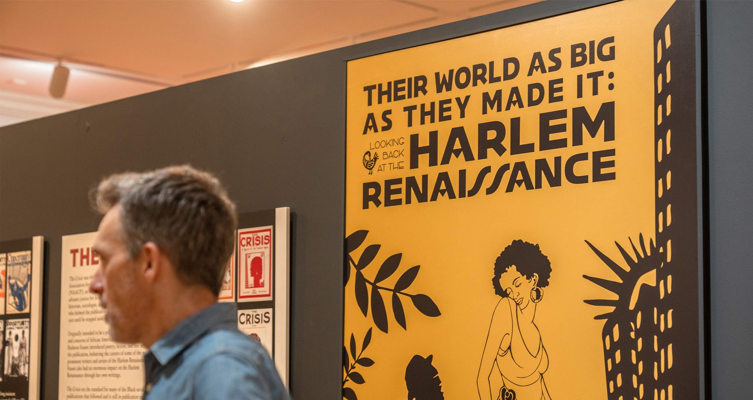 A man stands adjacent to a yellow sign with a woman on it that reads "Their World As Big As They Made It: Looking Back at the Harlem Renaissance."