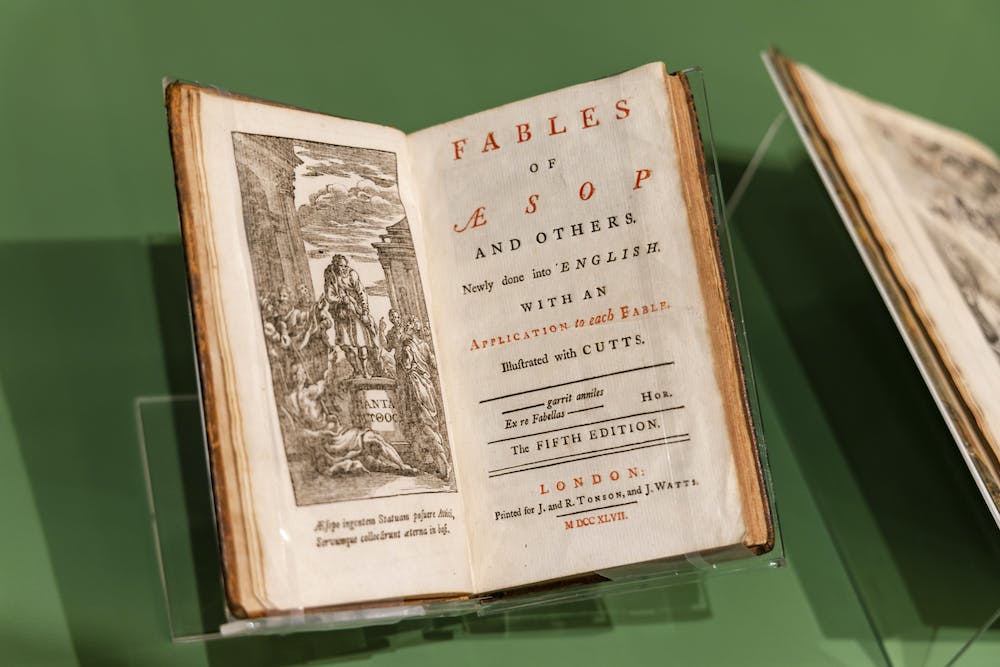 An old, faded book is propped open onto two pages: on the left, an illustration of a man standing on a pedestal; on the right, text that reads "Fables of Aesop and others. Newly done into English with an application to each fable. Illustrated with CUTTS. garrit anniles Ex re Fabellas — hor. The Fifth Edition. LONDON: Printed for J. and R. Tonson, and J. Watts."