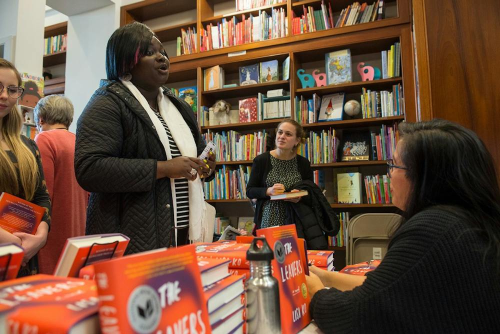 A woman sits in front of many stacks of books, whose covers read "The Leavers." She chats with another woman who stands in front of her.
