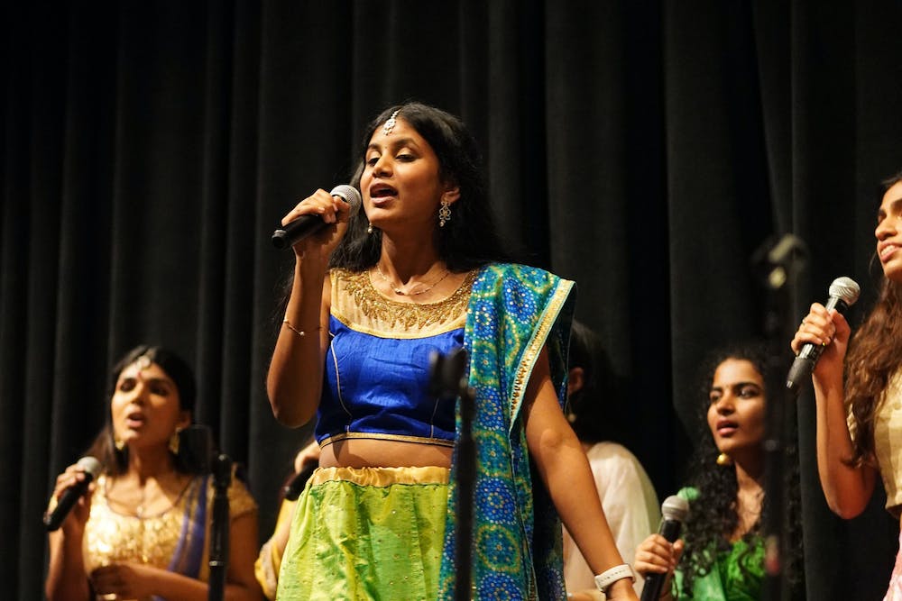 A woman dressed in blue, green and gold sings into a microphone, surrounded by others behind her also holding microphones.