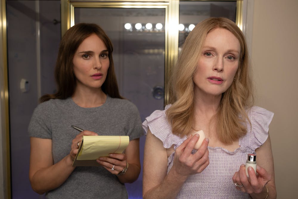 Natalie Portman, wearing a gray shirt and holding a pen and paper, and Julianne Moore, who holds makeup and a beauty sponge, look into a camera.