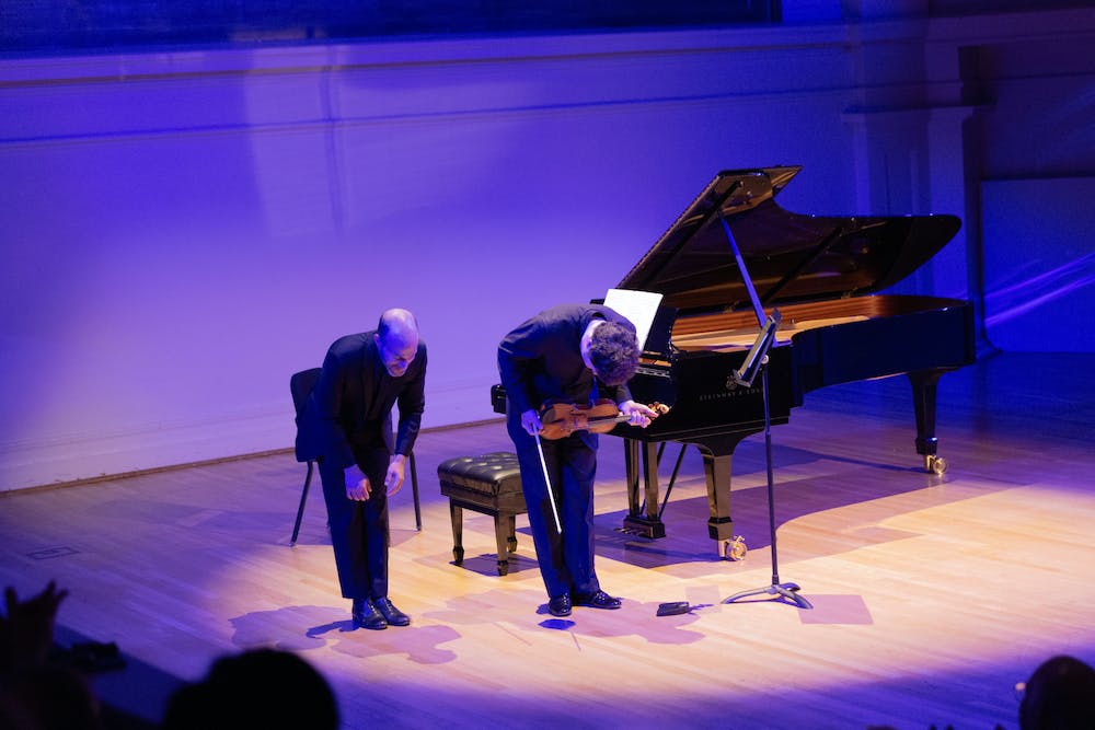 Hadelich and Weiss bow to the audience on stage at Old Cabell Hall, under a spotlight and next to a piano.