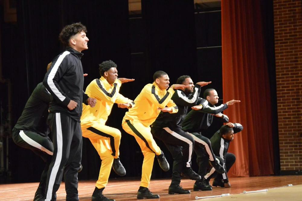 Seven men in black or yellow track suits perform a dance onstage, making their bodies look like the shape of a cobra.