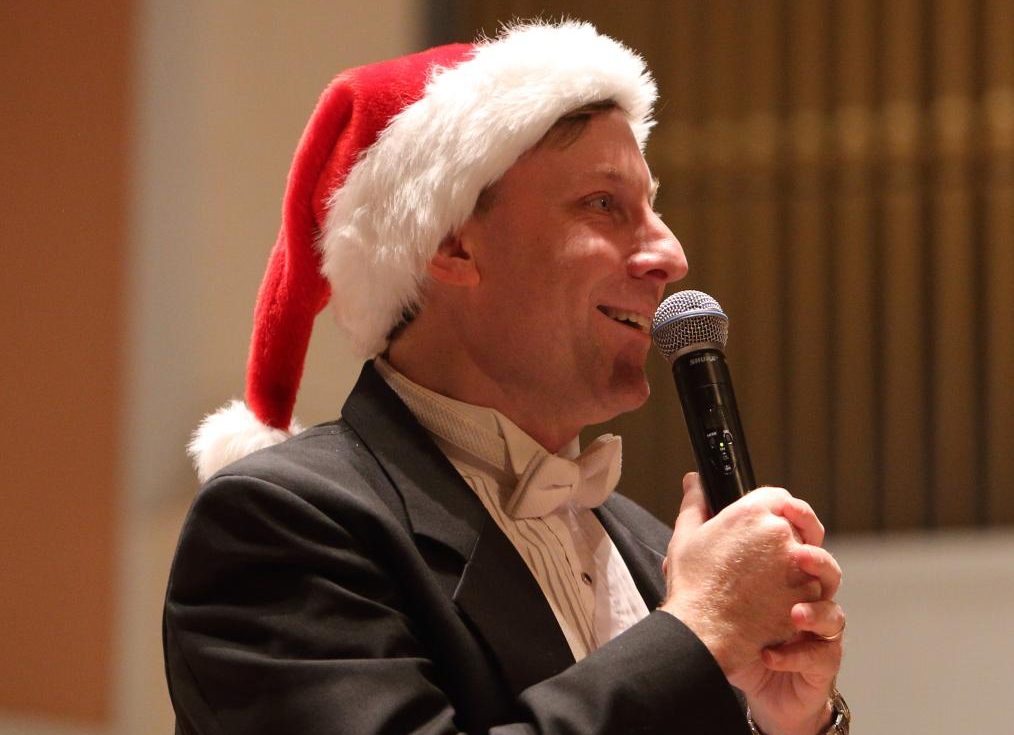 Michael Slon wears a tuxedo and a santa hat while smiling and singing or speaking into a microphone