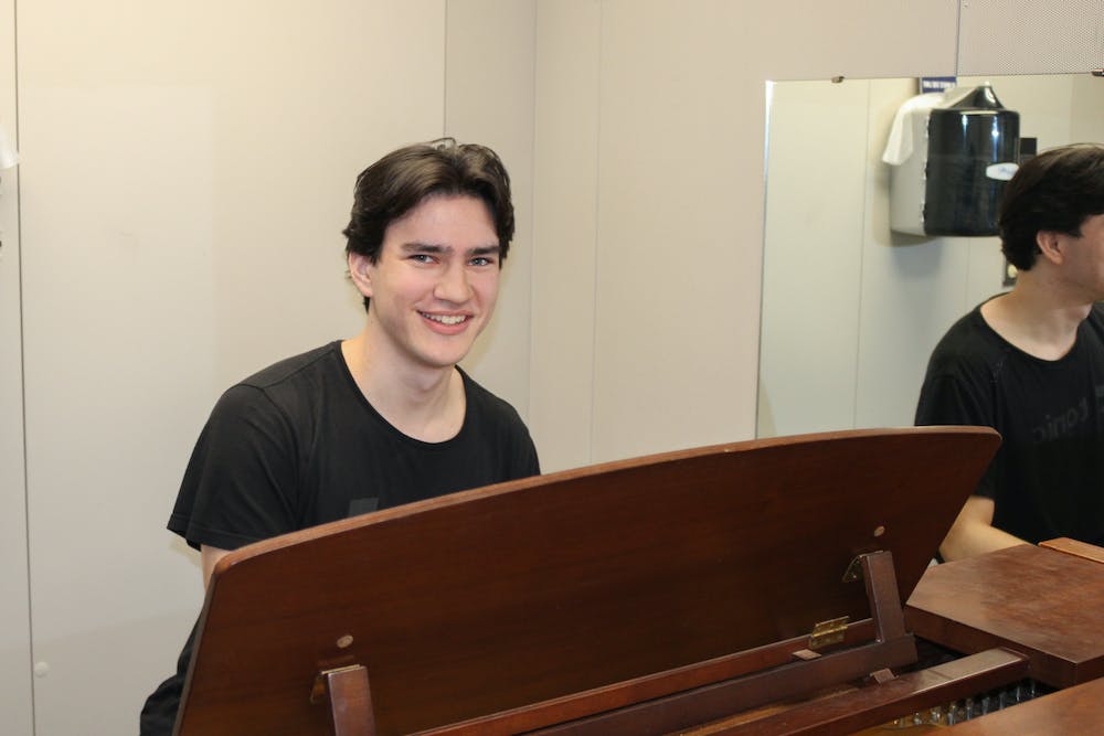 Fourth-year Piergiorgio Wilson sits at a piano and smiles for the camera next to a mirror.