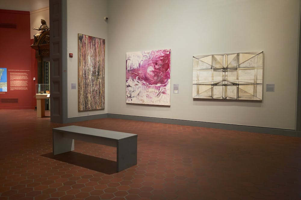 Three abstract pieces hang on the wall of a room in the Fralin Museum. From left to right: (1) features multicolored, drip-like streaks; (2) a white canvas with pink and magenta swirls, (3) a horizontal brown and white piece made up of geometric shapes