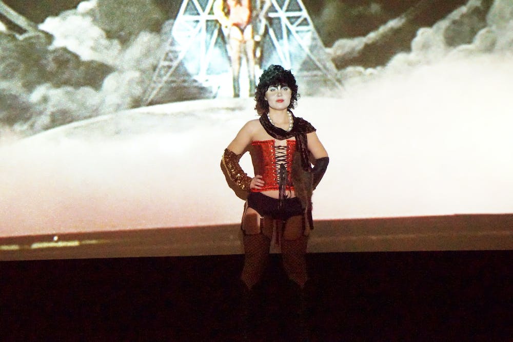 A person with black curly hair, heavy theatrical makeup, and a red corset stands with their hands on their hips in front of a projected screen. 