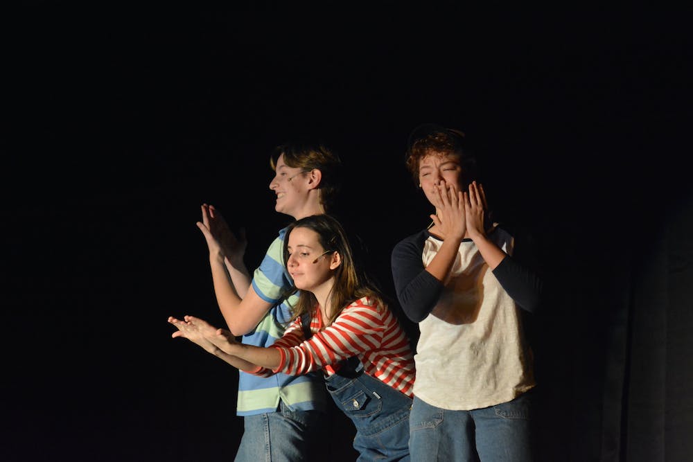 Students Levi Green (wearing a blue and green striped shirt), Bridget Gauntner (in overalls and a red-and-white striped tee), and Laila Bolte (in a white and blue ringer tee) hold their hands up and sing to the audience.