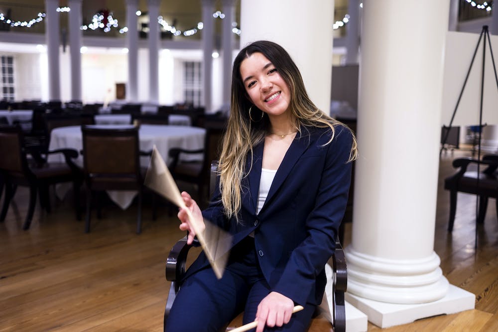 Emma Heraldo, dressed in a blue blazer and dress pants, sits in the rotunda and twirls a drumstick in hand, beaming at the camera.
