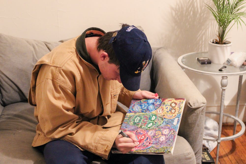 Holden Brown, wearing a brown jacket and a navy blue baseball cap, draws faces on a colourful canvas.