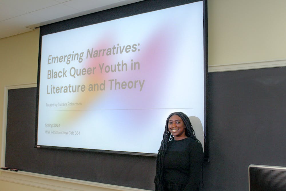 Tichara Robertson wears a black shirt and stands in front of a projector that reads, "Emerging Narratives: Black Queer Youth in Literature and Theory"