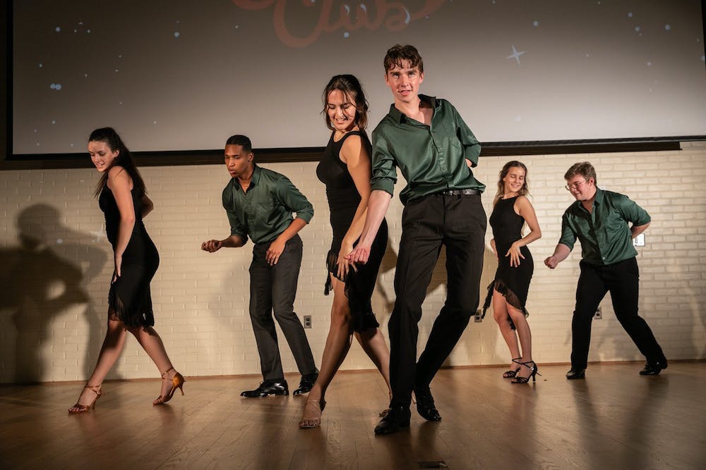 A group of students perform a salsa number on a stage. Some performers wear black dresses, and others wear black pants and green shirts.