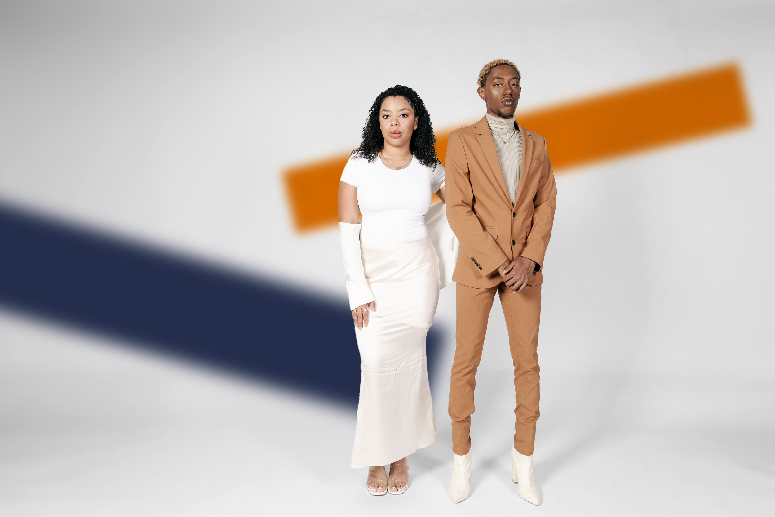 Two students pose in front of a white background with a blue stripe and an orange stripe. One wears a white t-shirt, a long beige skirt and white fingerless gloves; the other wears a tan suit and a beige turtleneck. 
