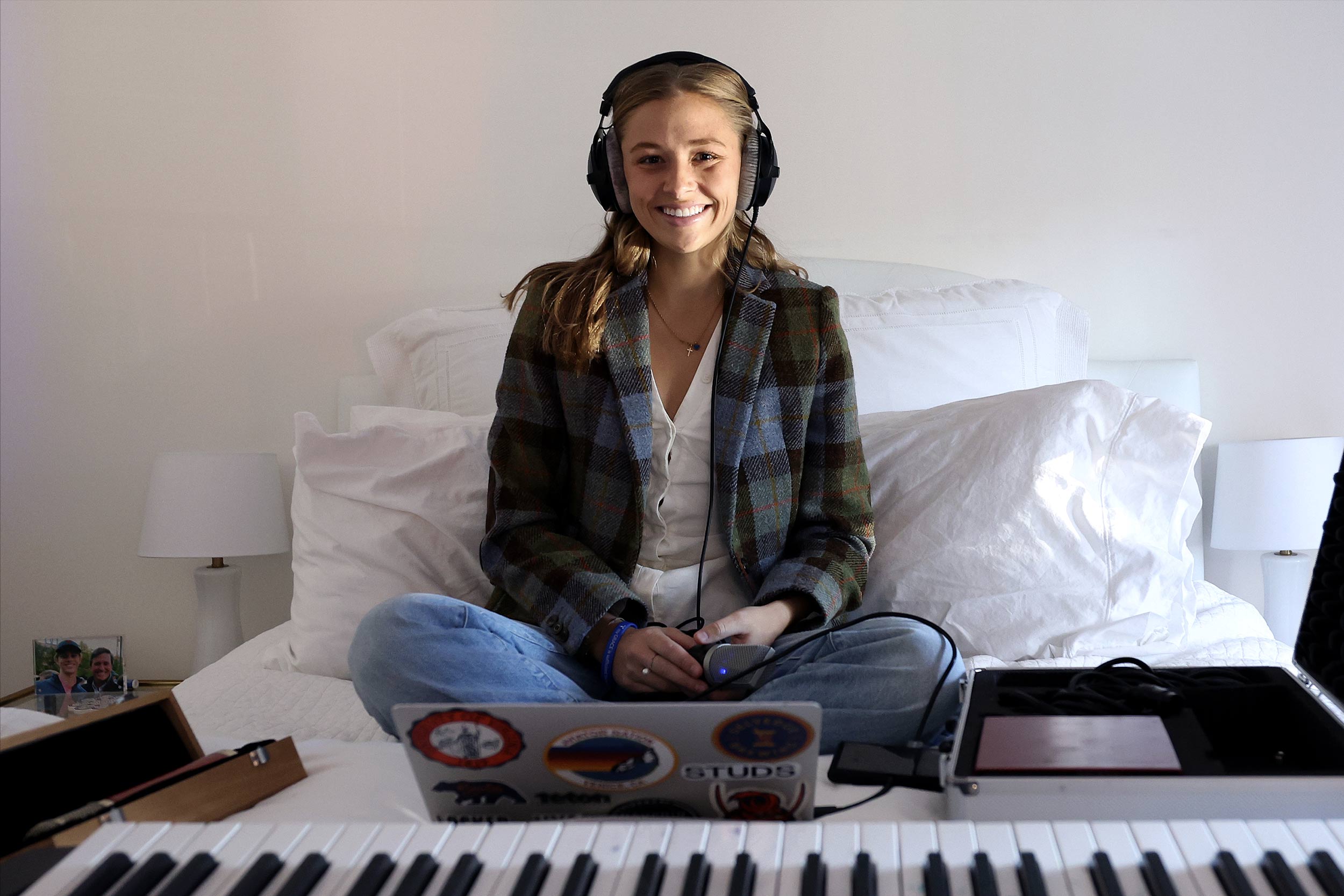 Claire Frazier sits cross-legged on a bed in front of a keyboard and a laptop, wearing headphones and a plaid jacket. 
