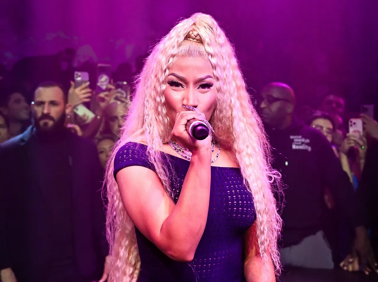 Nicki Minaj, wearing a long, crimped blonde ponytail, holds a microphone to her mouth and looks at the camera while bathed in pink-purple light. 
