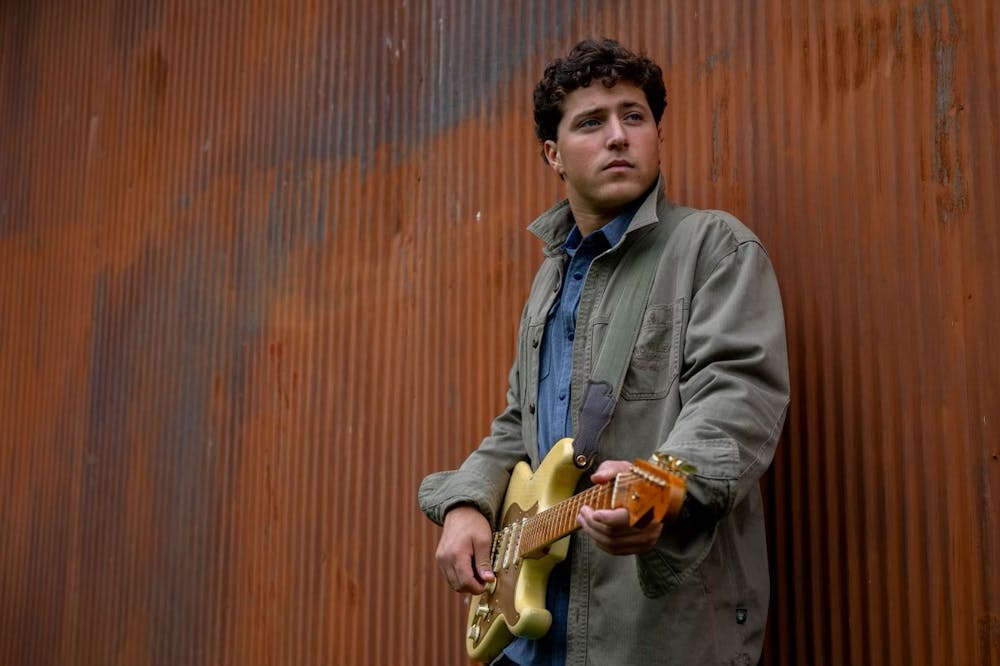 Jack Stepanian holds an electric guitar while staring off into the distance, leaning against a rusted orange wall.