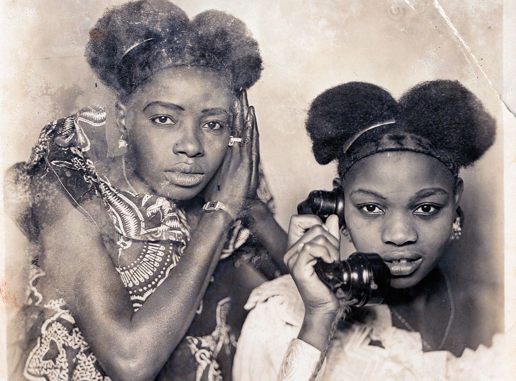 Two women pose for a photo taken in a studio in the 1950s — one holds her hands next to her face while staring at the camera, while the other holds a telephone to her ear.