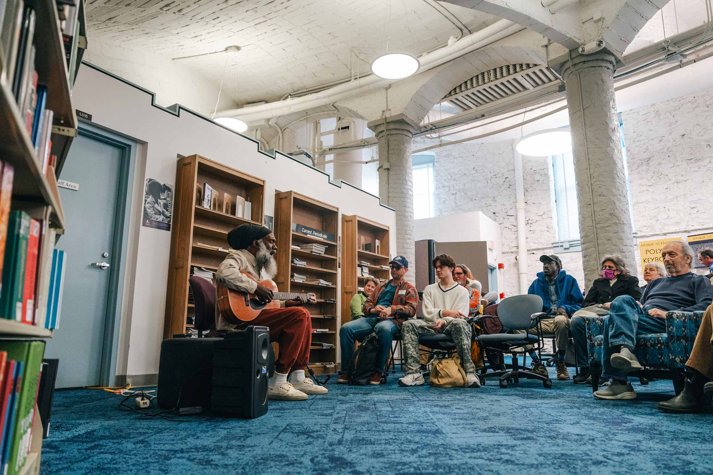 Musician Corey Harris sits on a chair strumming a guitar in the music library, which has high white walls and ceilings and a blue carpet. He is watched by an audience, also sitting in chairs.