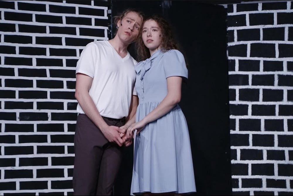 Performers Soren Corbett and Laila Bolté, playing characters Alfred Hallam and Alice Spencer, join hands and look out into the audience in front of a black-and-white brick background.