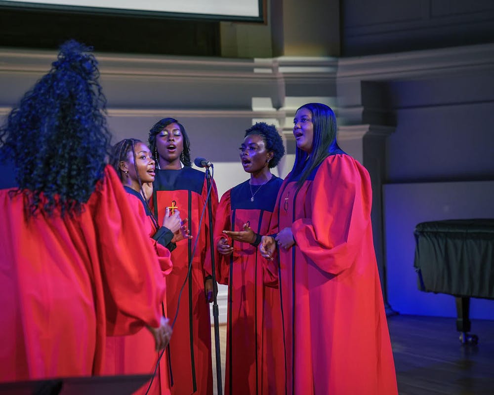Four gospel singers wear red robes and stand around a microphone and sing. Another member stands with her back to the camera. 