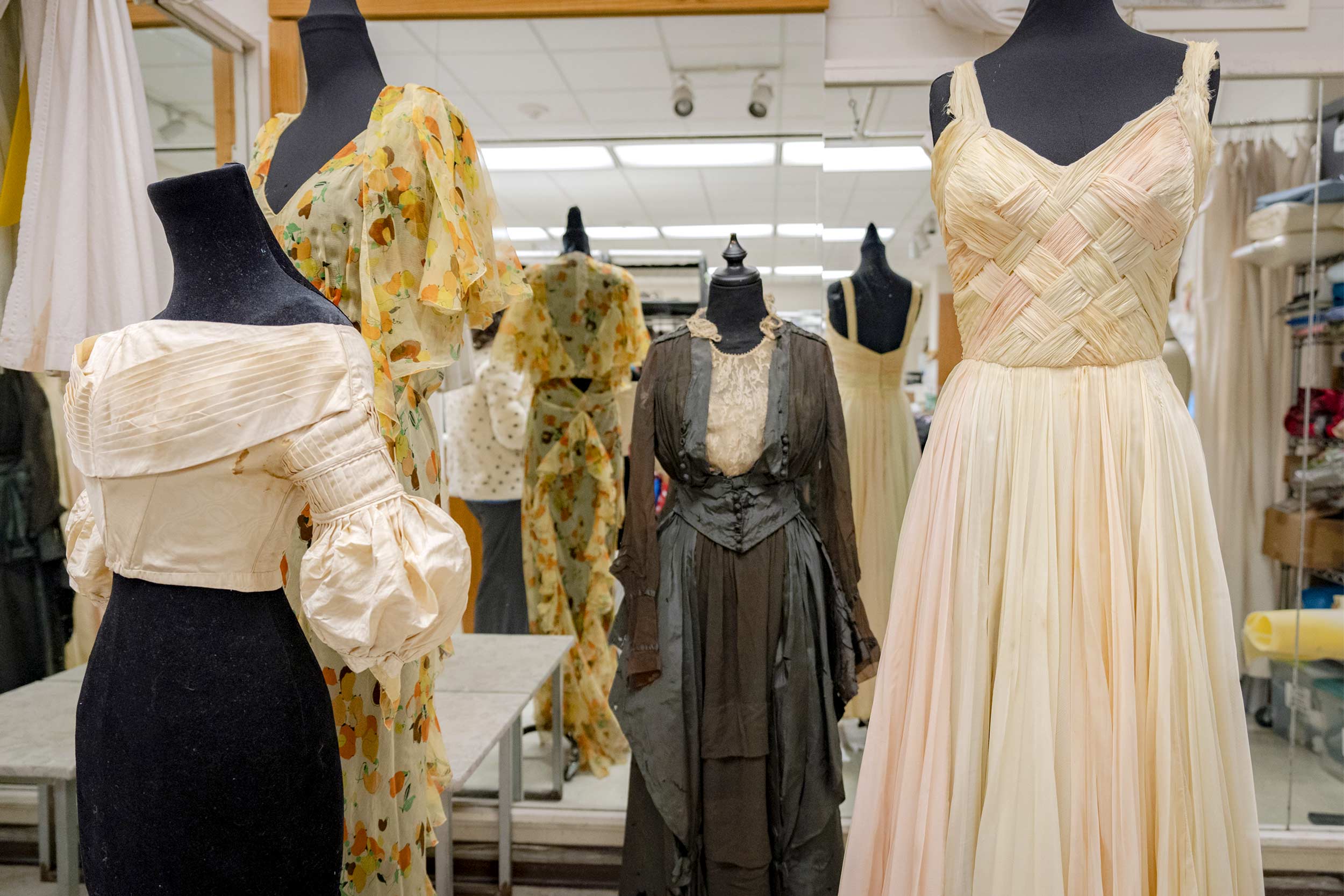 Several dresses are shown on mannequins. One on the left features an intricate top with puffy sleeves and a plain black skirt; another towards the back features flowy sleeves and yellow, orange and brown fruit; an antique black and white dress; and an off-white sleeveless dress with a lattice pattern.