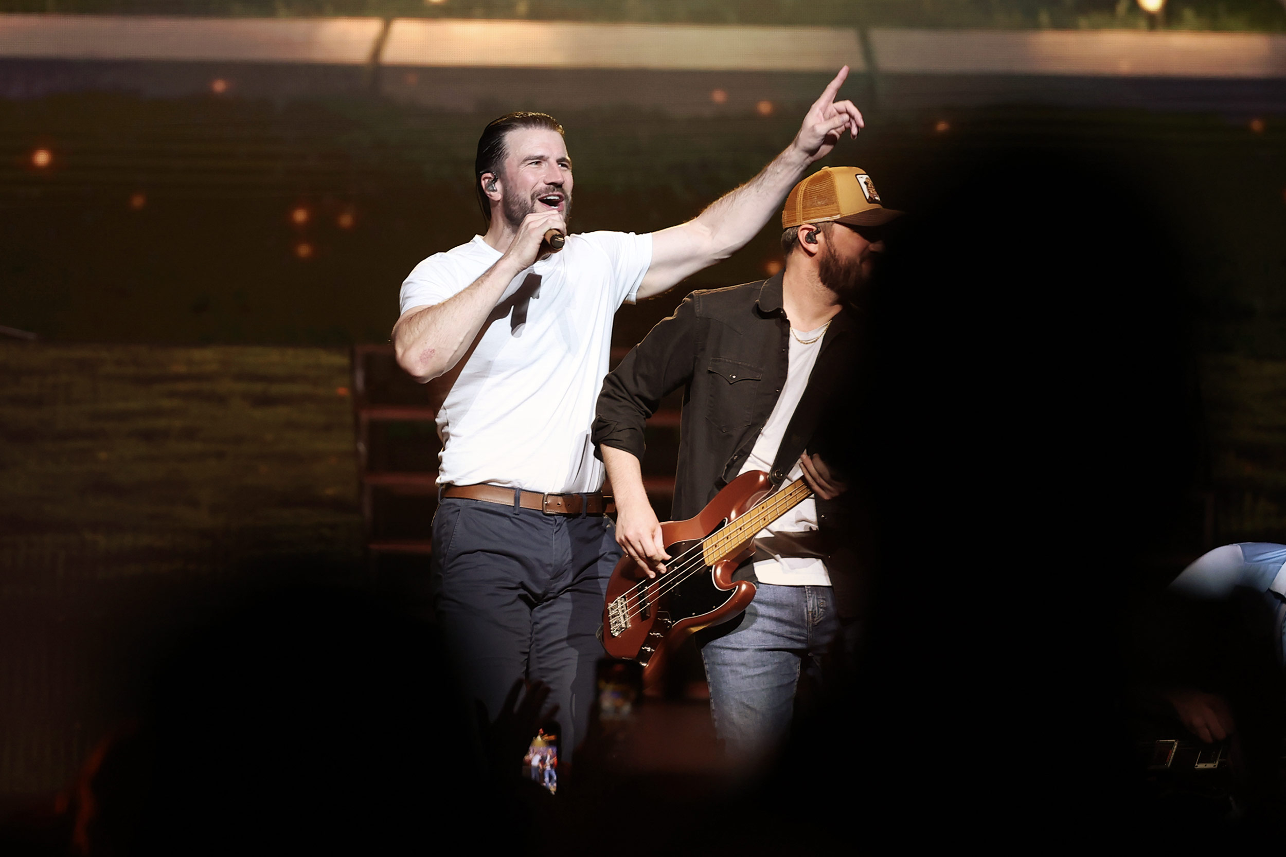 Sam Hunt smiles onstage wearing a white shirt and dark blue pants while holding a microphone. In the back, a man wearing a cap plays the guitar.