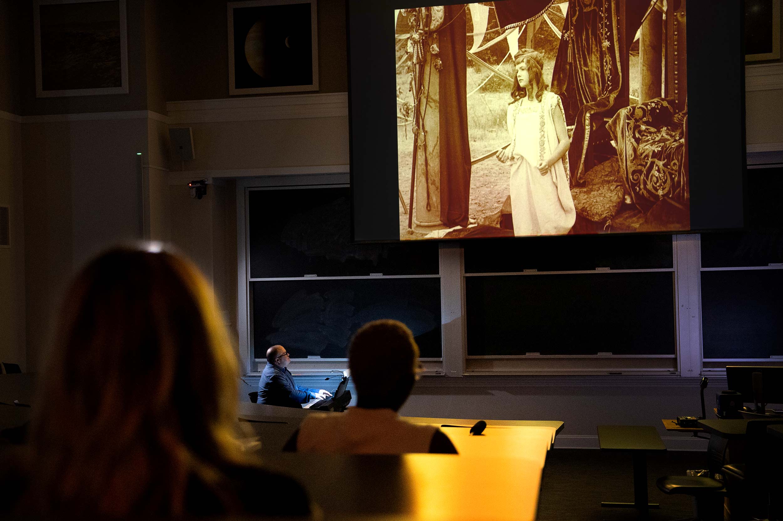 Audiences sit in a classroom facing a projector screen, which shows a sepia-toned scene featuring a girl in a dress. Matthew Marshall sits at a piano under the projector screen.