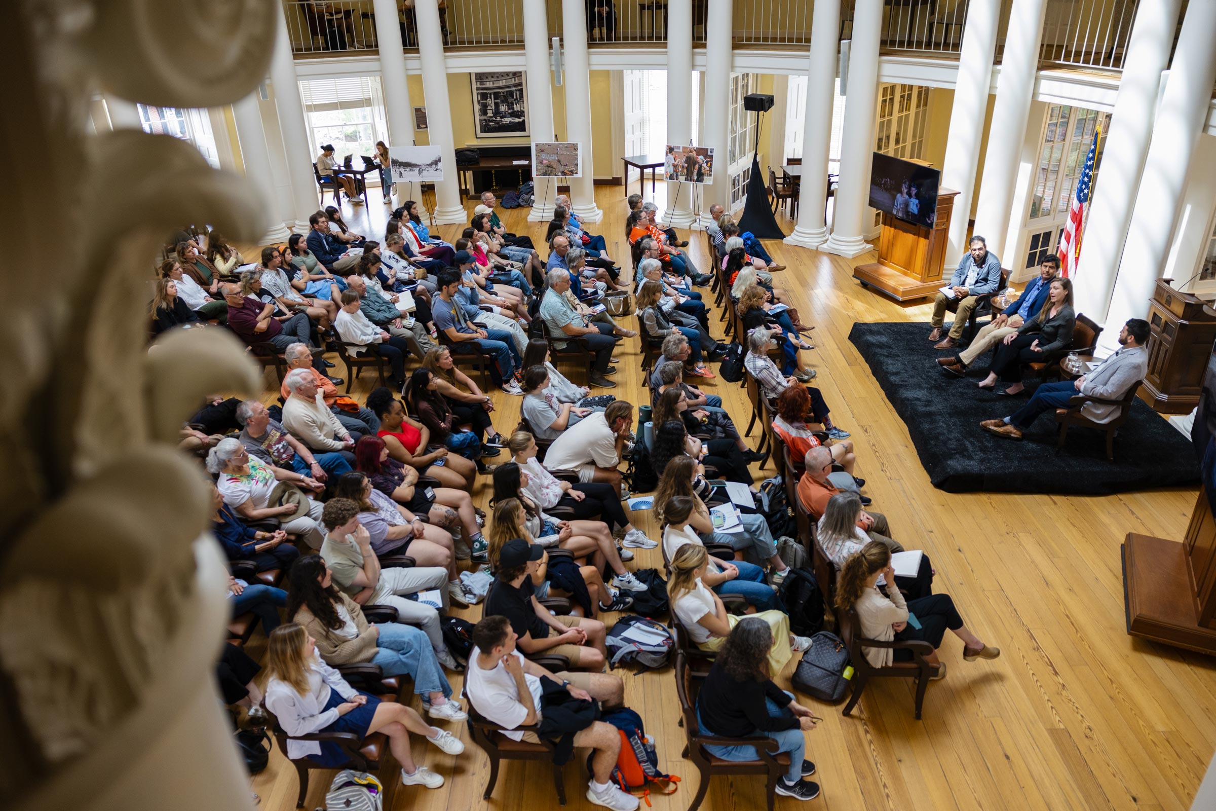 A large audience sits in rows of chairs inside the Dome Room of the Rotunda, facing a small stage of four speakers.