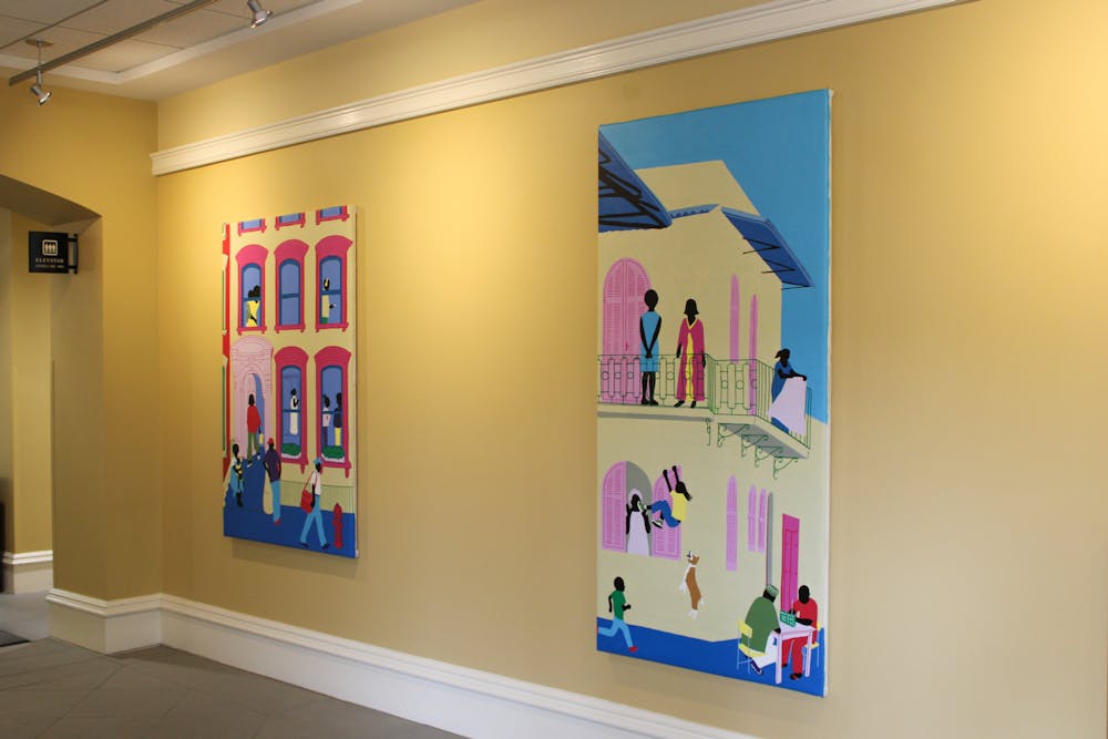 A yellow wall holds two paintings by artist Uzo Njoku — both paintings feature pastel renderings of buildings that feature windows and faceless figures existing in and around them.