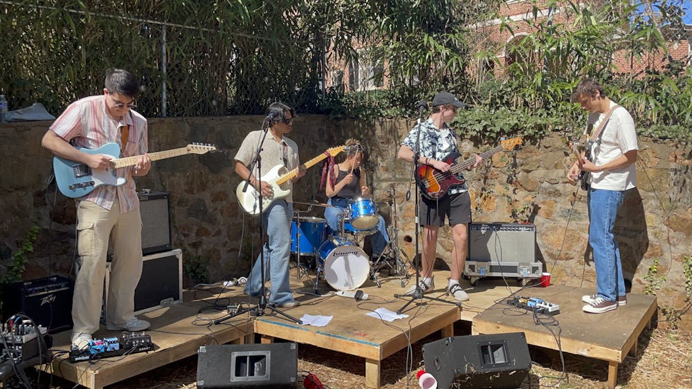 Five members of the band no composure play guitars, a bass, and a drum set on a sunny day on top of a wooden platform.