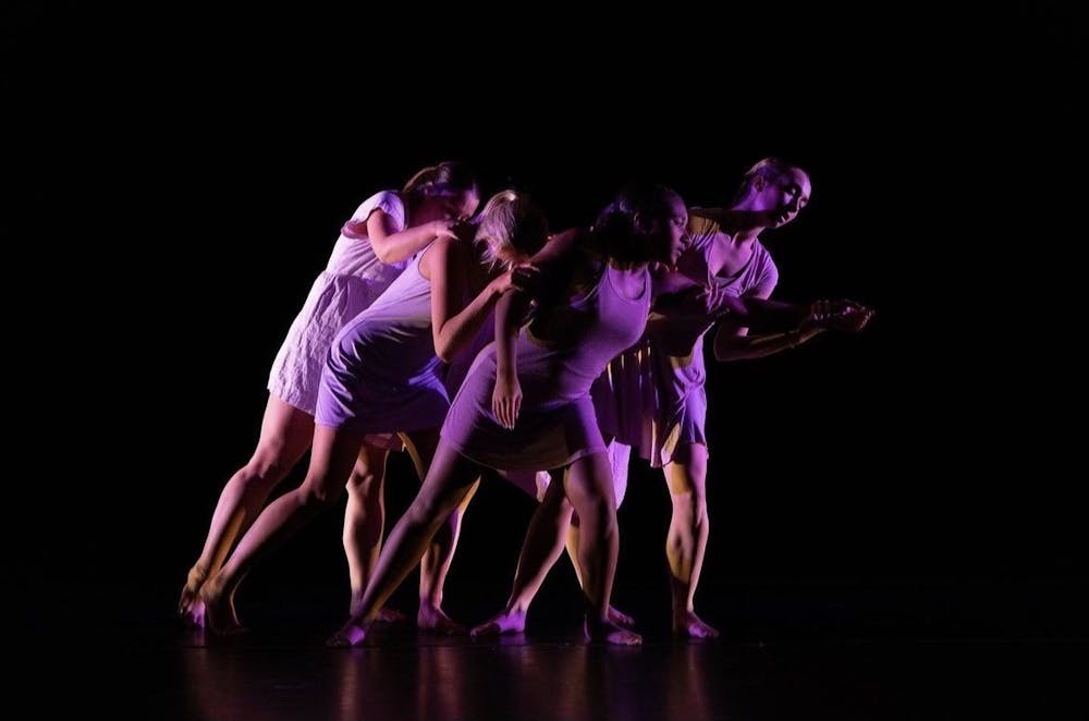 A group of students dressed in neutral colors perform a contemporary dance under pink and purple lighting. 