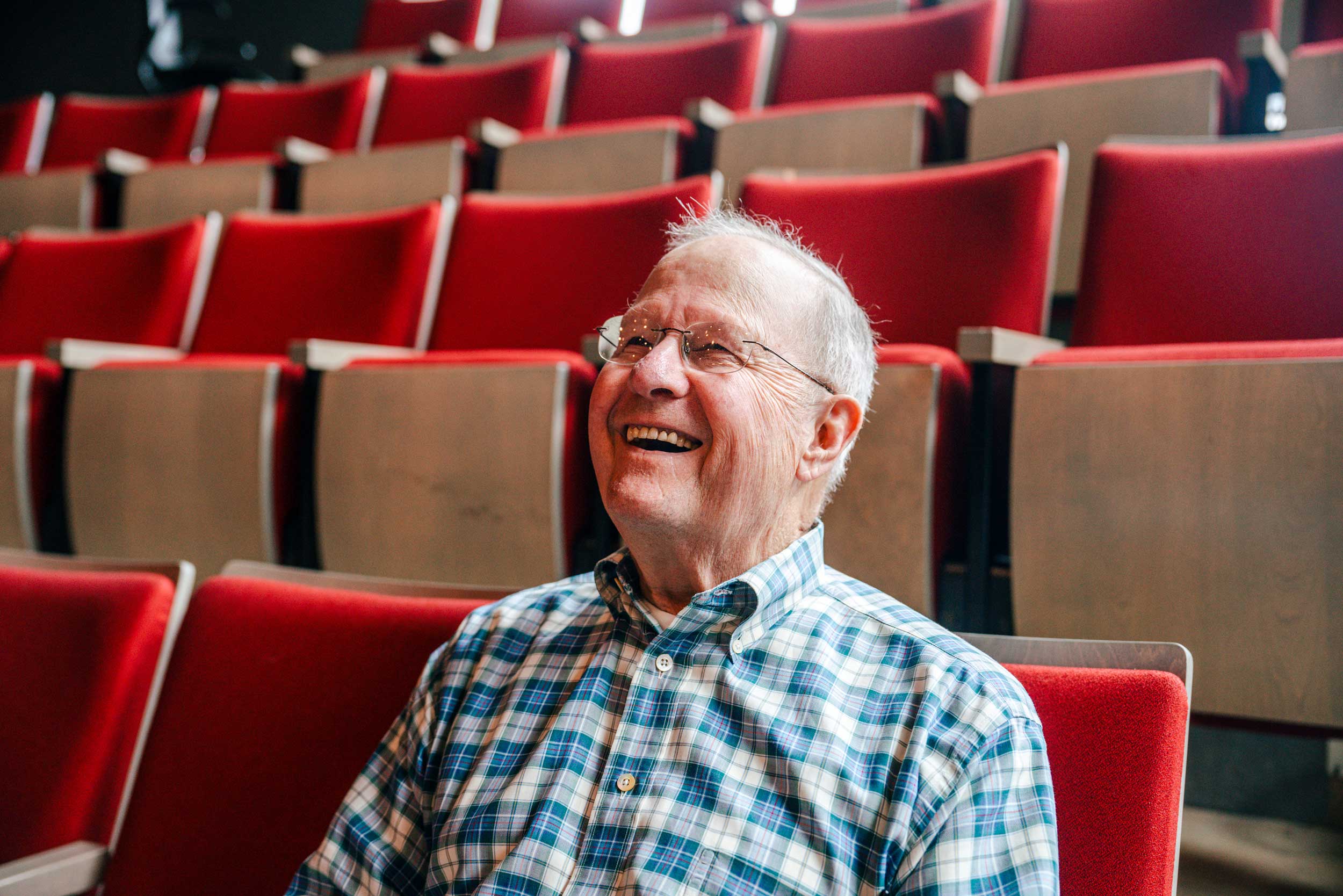 Drama professor emeritus Bob Chapel sits in an empty theatre with red chairs, smiling. 