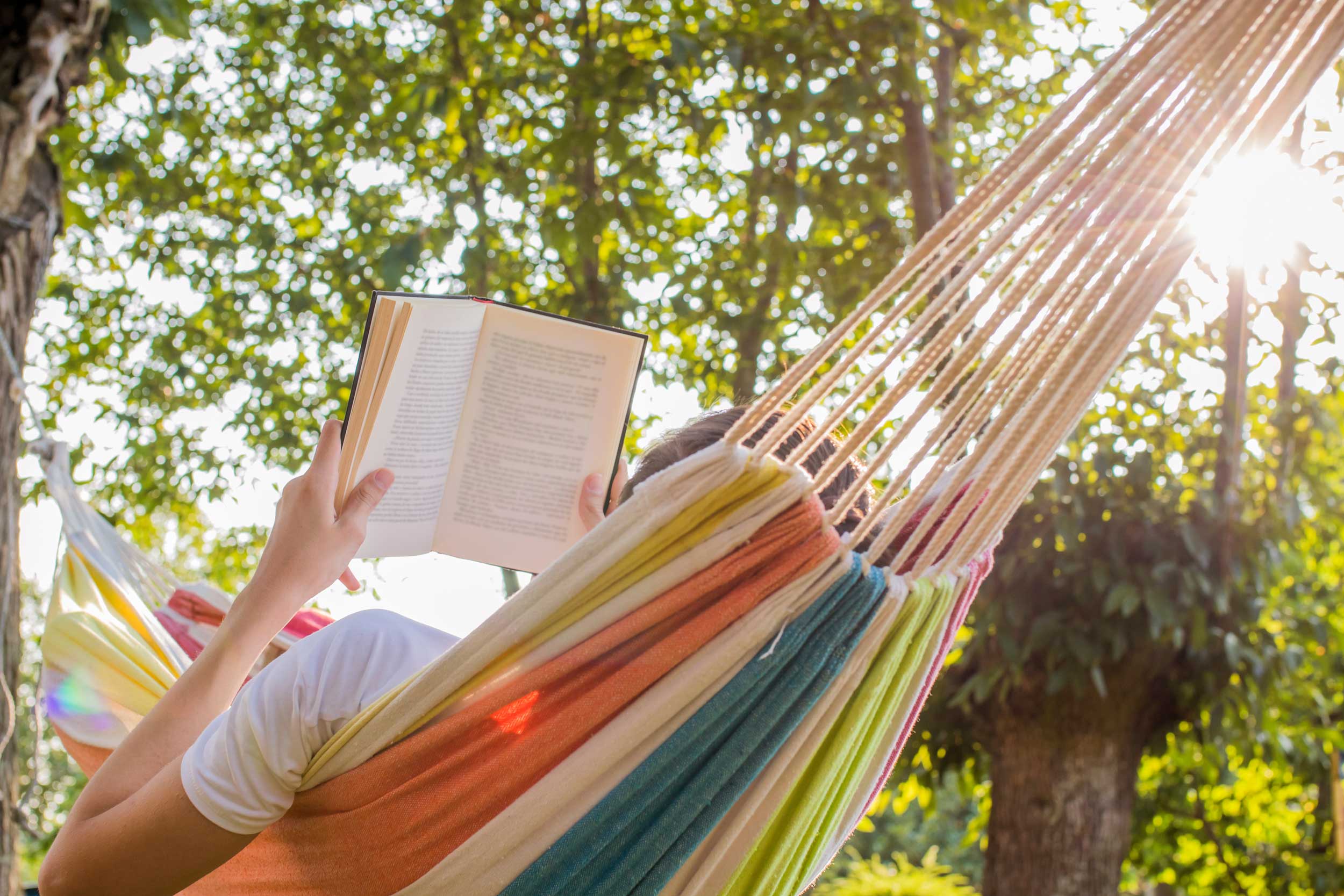 A person lays in the sun with an open book on a colorful hammock.