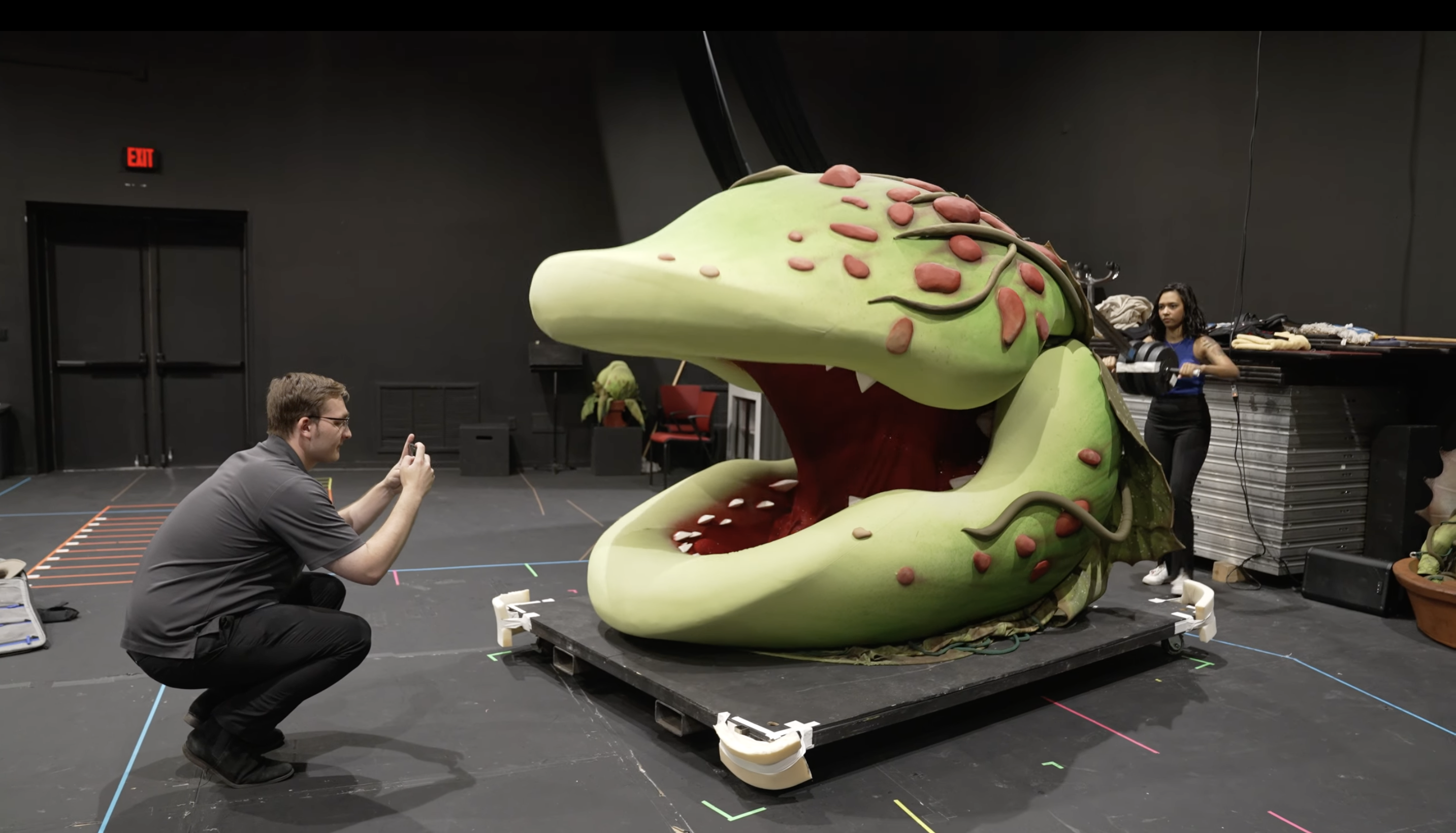 An actor operates Audrey II, a giant puppet of a green plant with a mouth and teeth, opening its mouth.