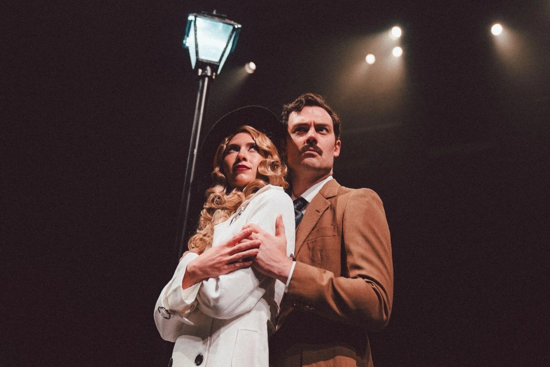 Woodrow Proctor as Richard Hannay embraces Mollie Downes as Annabella Schmidt. They look into the distance under a prop streetlight and stage lights.