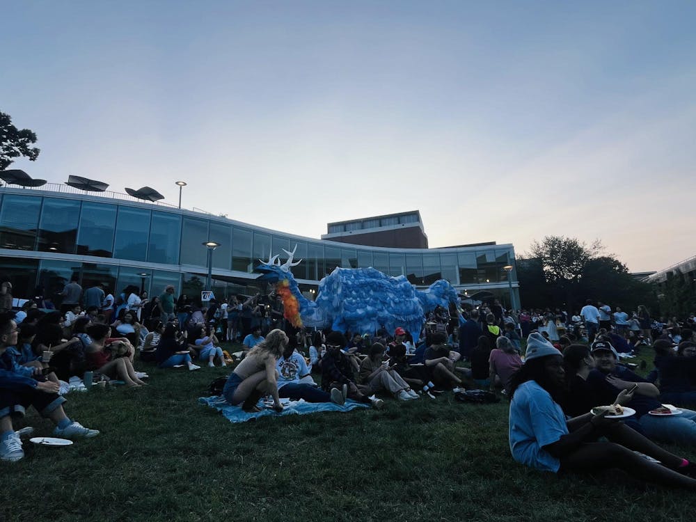 Many people sit on the ground outside of the Drama Education Building. A giant blue and orange puppet is paraded across the lawn.