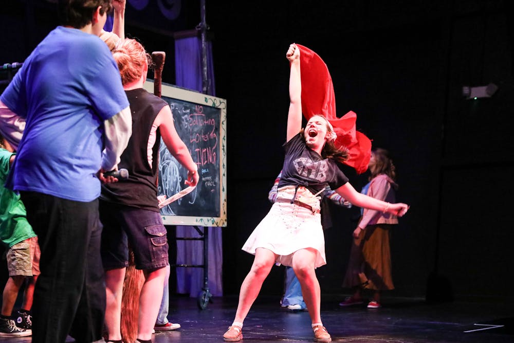 A performer stands onstage in a black t-shirt and a white skirt, raising a red flag above their head and smiling wide with their eyes closed. Two other performers look on the first one, their backs to the camera.