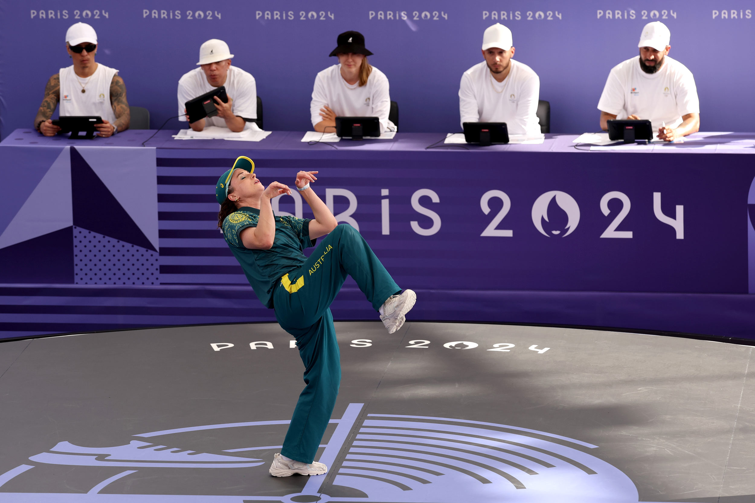 Australian dancer Raygun dances in a green jumpsuit in front of four judges wearing white on a purple panel that reads "PARIS 2024".