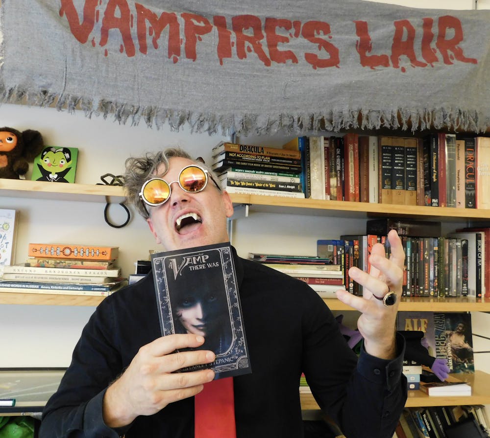 Professor Stanley Stepanic wears fake vampire teeth and circular sunglasses while holding up his book, titled "A Vamp There Was." The cover is black and includes a shadowy photo of a woman smirking and looking at the reader.