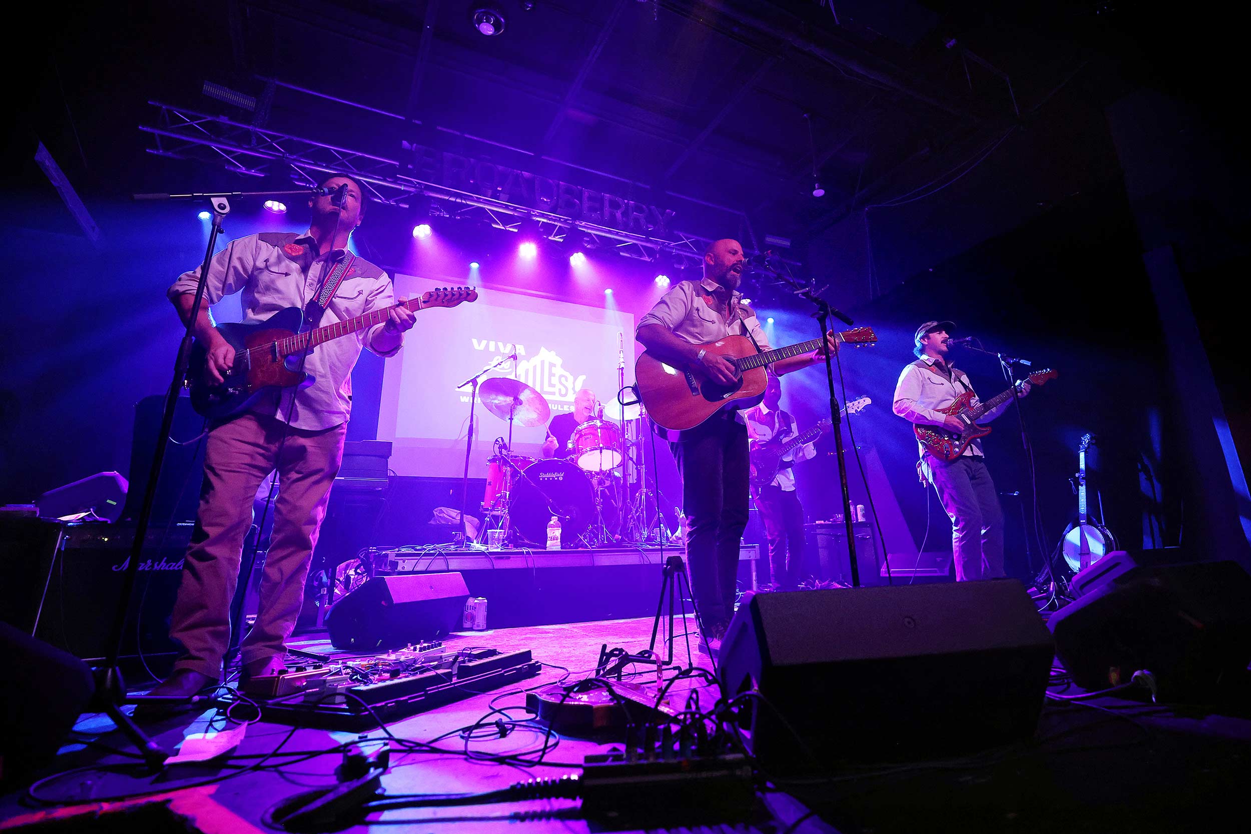 Band members Mason Brent, Andy Stepanian and Jack Heard stand on a stage lit by purple lights and play guitars.