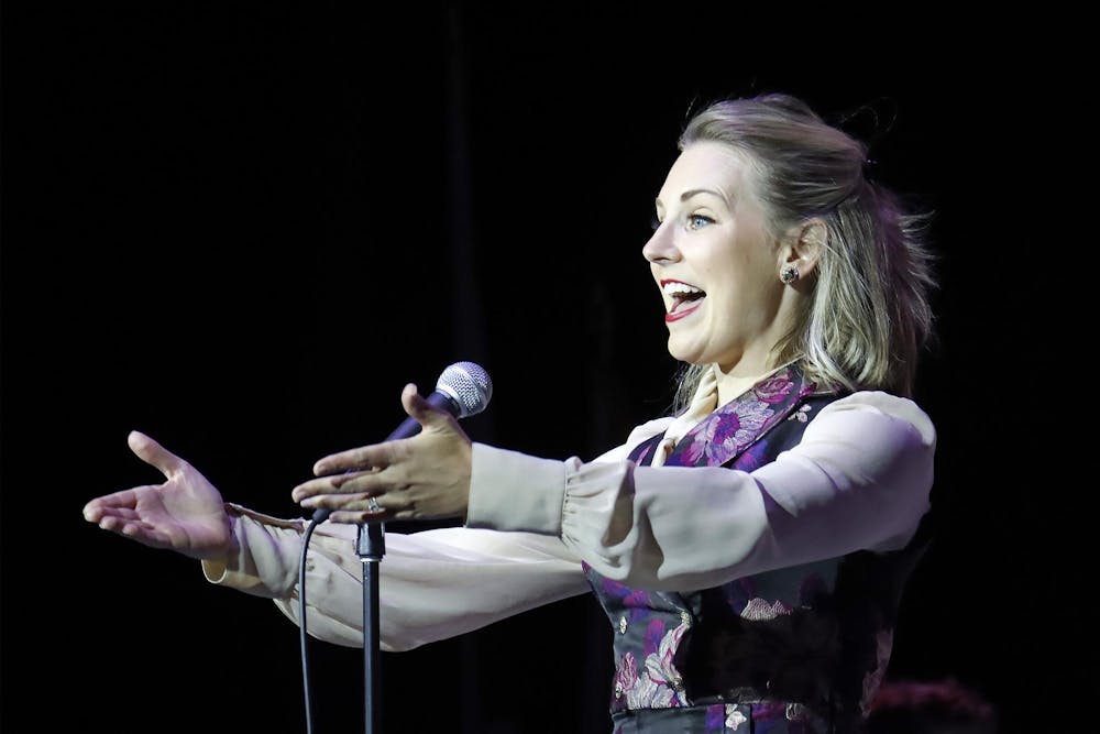 Jenna Pastuszek is shown singing into a standing microphone with her arms outstretched in front of her.