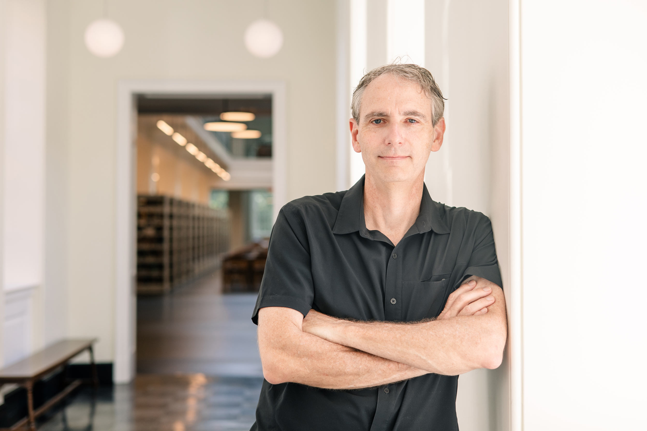 Bruce Holsinger leans against a white wall with his arms crossed, wearing a dark gray collared shirt, smiling at the camera. A hallway in Shannon Library is seen in the background.