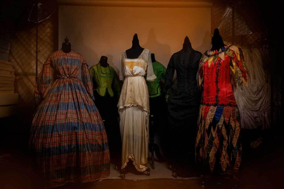 A gallery room showcases six historical outfits displayed close together in mannequins. There is dark, moody lighting. 