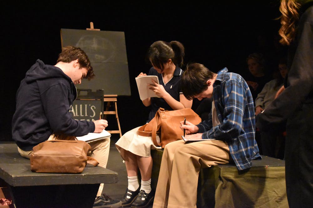 Three students sit in a black box theatre on benches, writing in their script books, with a black chalk board on an easel behind them.