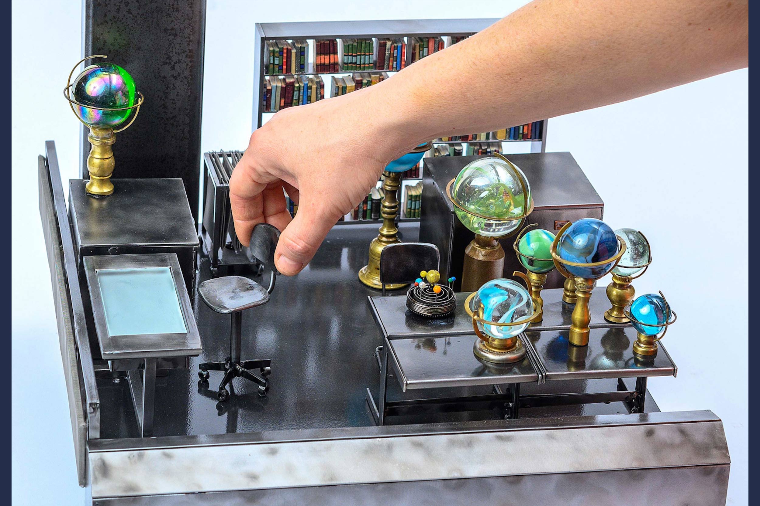 A close up image of a hand adjusting a small chair in a miniature display. The display shows a room with various desks and rolling chairs, and globes placed on the desks. One of the walls is a large bookshelf, filled entirely with colorful books. 